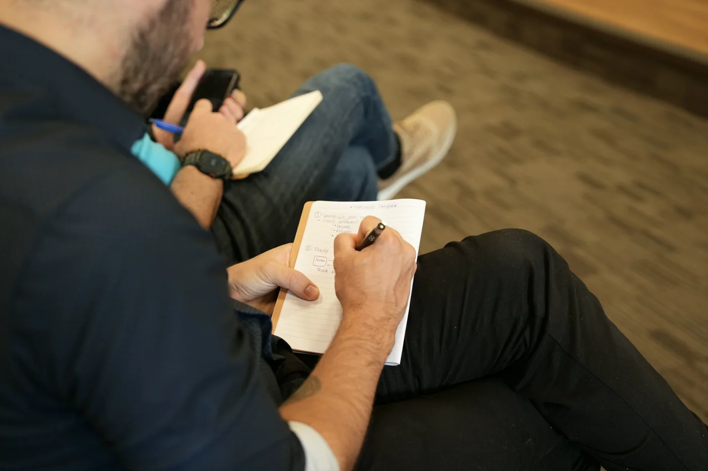 Mão anotando à caneta em um caderno pequeno, apoiado sobre o joelho, com um celular na outra mão.